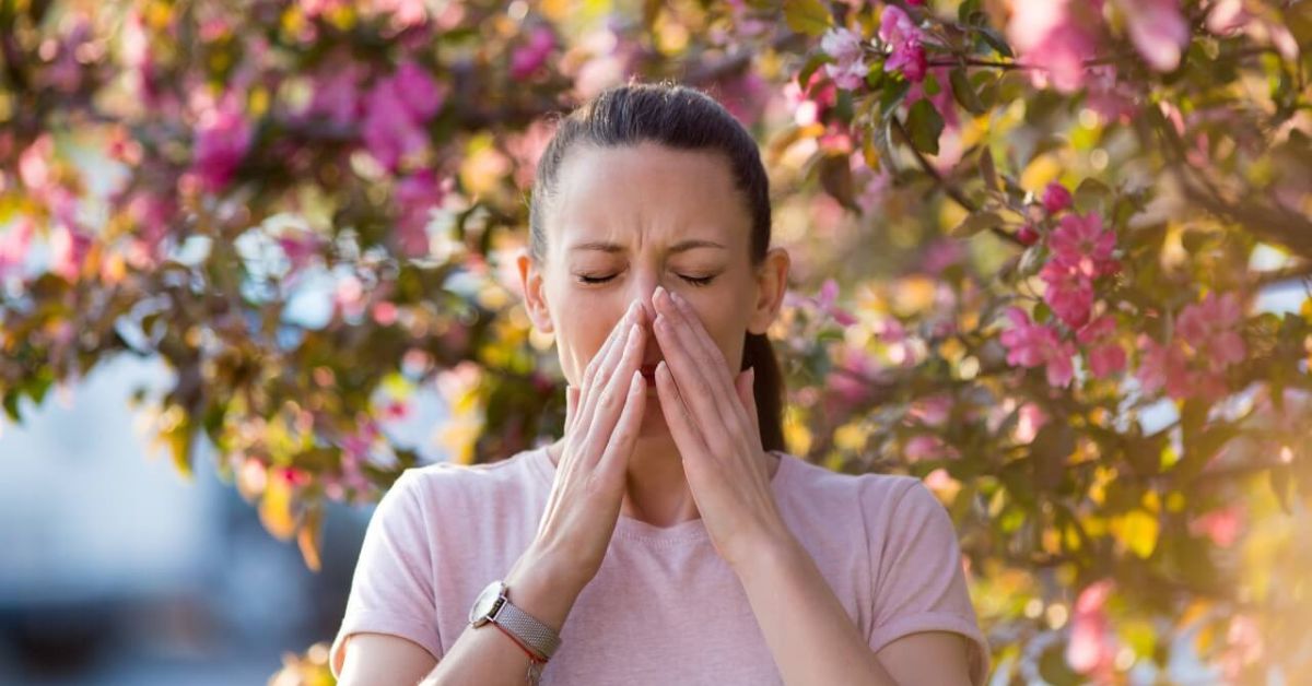 uma mulher próximo a uma arvore com flores cor de rosa, com as mãos no nariz espirrando devido as alergias de primavera