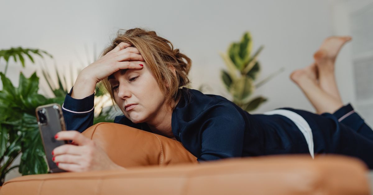 Mulher loira com roupa azul, mão na cabeça deitada na cama, olhando para o celular parecendo estar com bad de domingo