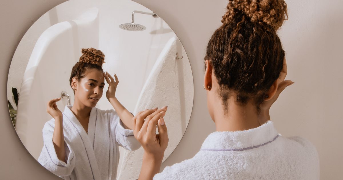 menina com um coque no cabelo, de roupão, se olhando no espelho, passando creme no rosto e realizando seu skincare
