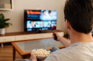 homem sentado, com balde de pipoca na mão e controle da televisão na outra, ligando a tv e evitando a bad de domingo